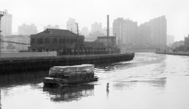A covered boat travels along a river beside industrial buildings, with modern high-rise structures visible in the misty background. The scene is in black and white.