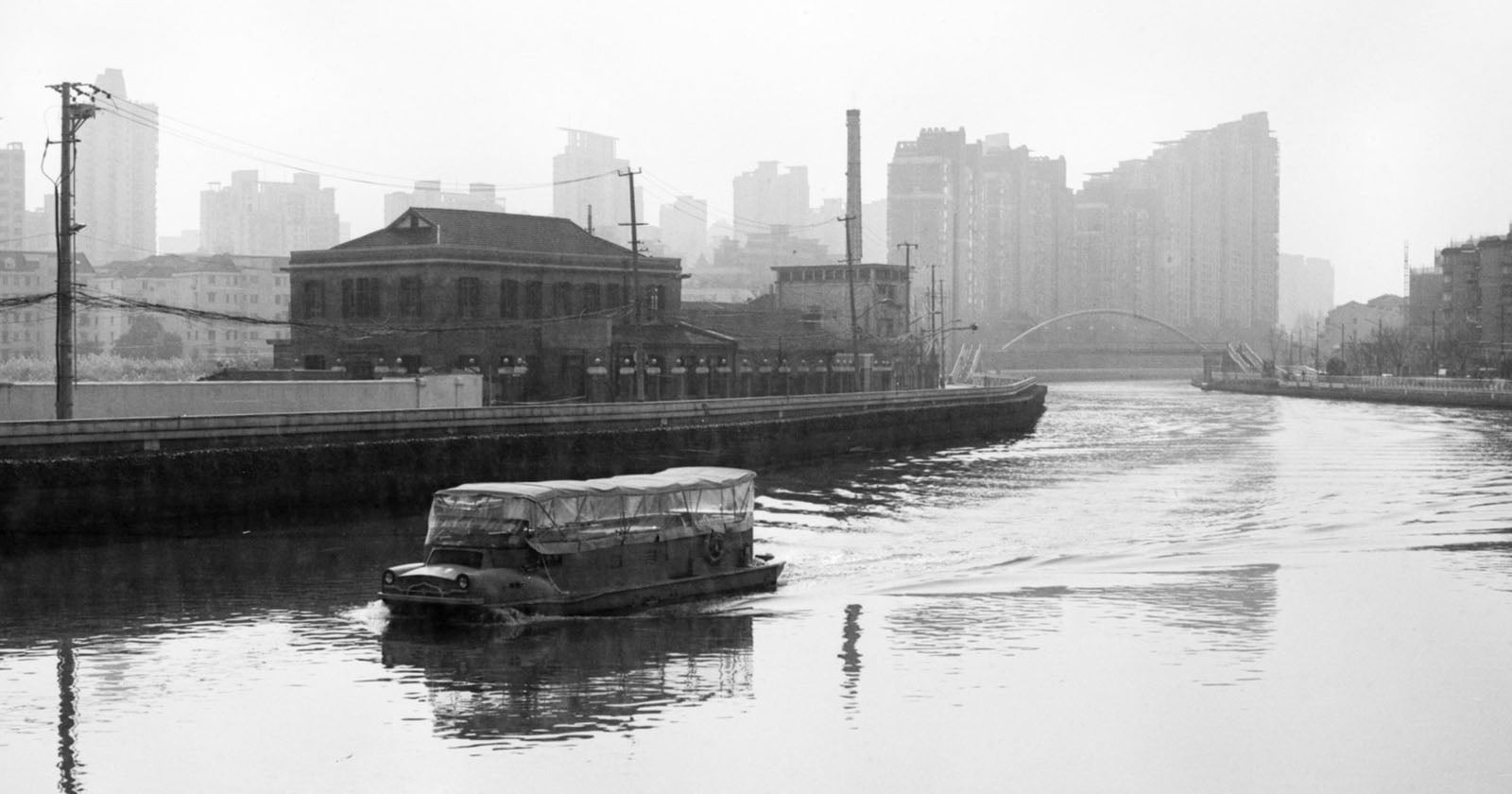 A covered boat travels along a river beside industrial buildings, with modern high-rise structures visible in the misty background. The scene is in black and white.