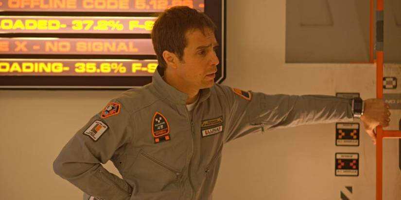 Sam Bell leaning and gripping a small red mini ladder in a white and gray room and looking to the right in Moon.