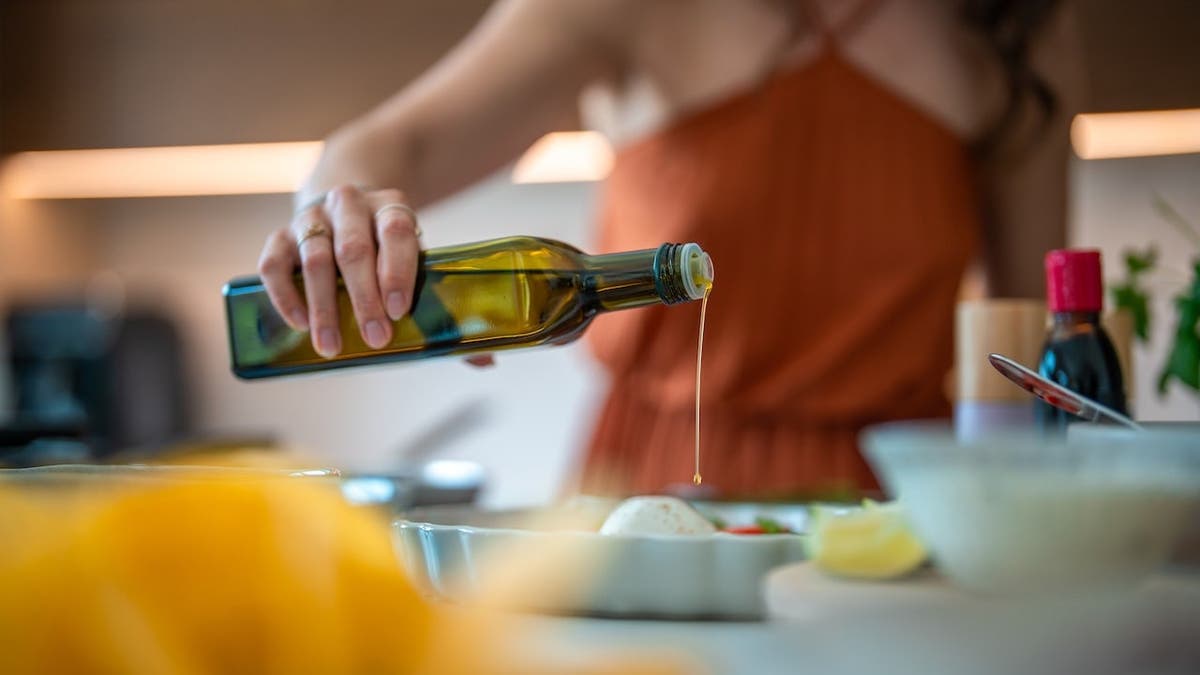 Feminine hands in orange apron pouring golden olive oil during meal preparation.