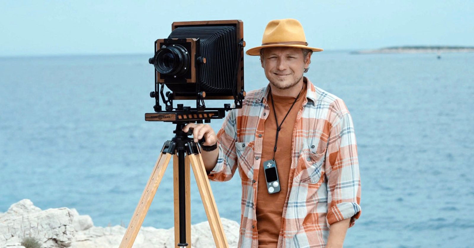 A man wearing a yellow hat and plaid shirt stands by the sea, smiling next to a vintage large-format camera mounted on a tripod. Rocky shore and calm blue water are in the background.