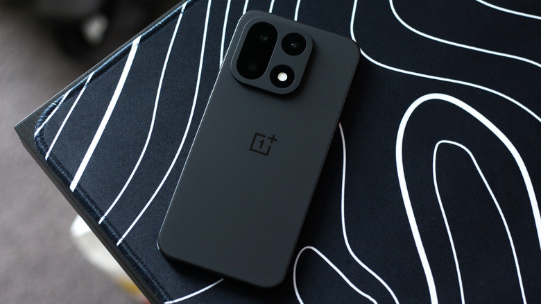 Black OnePlus 15 on a mousepad with an abstract white and black pattern.