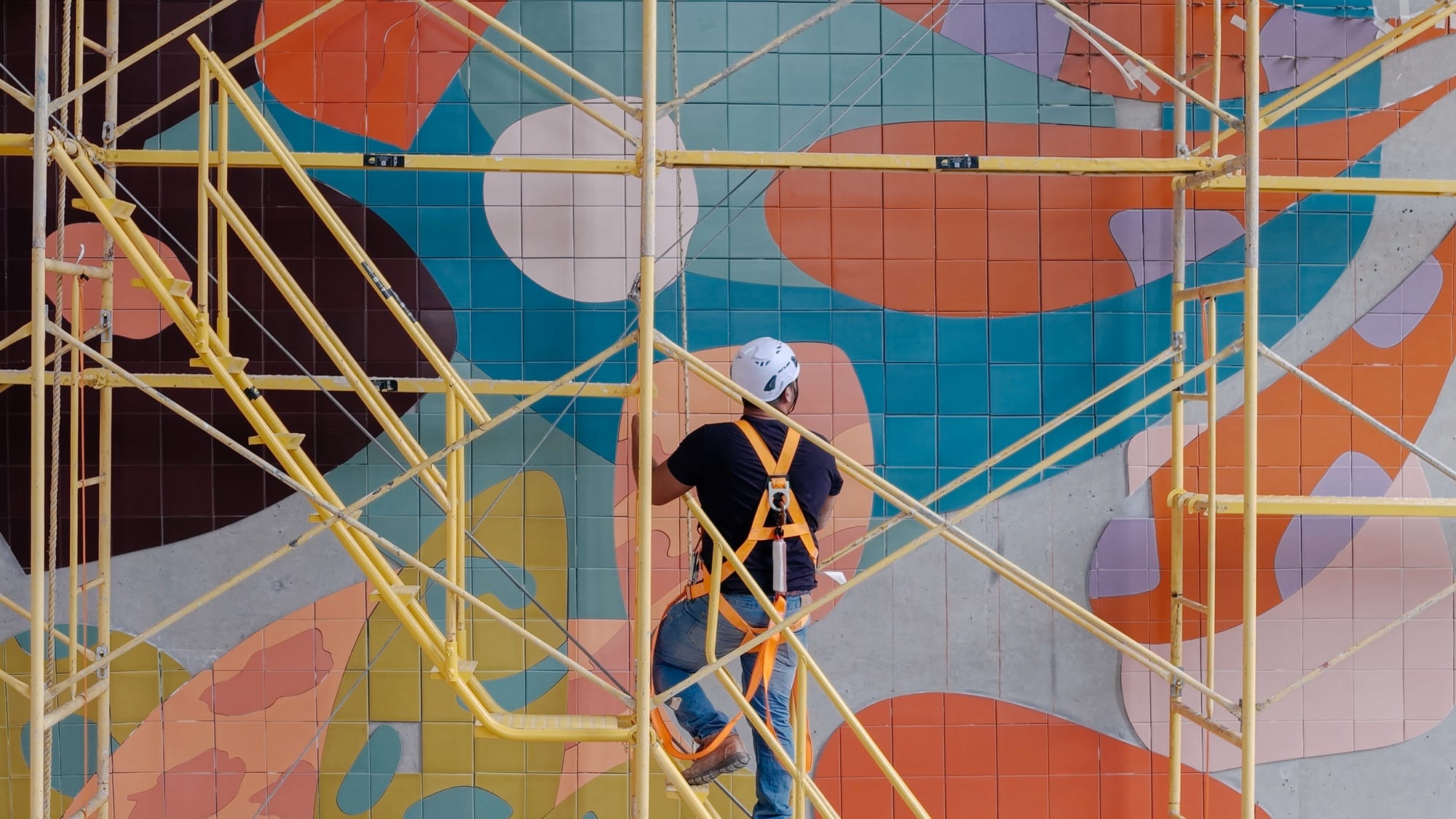 a person works on a mural by Alex Proba of colorful ceramic tiles amid scaffolding