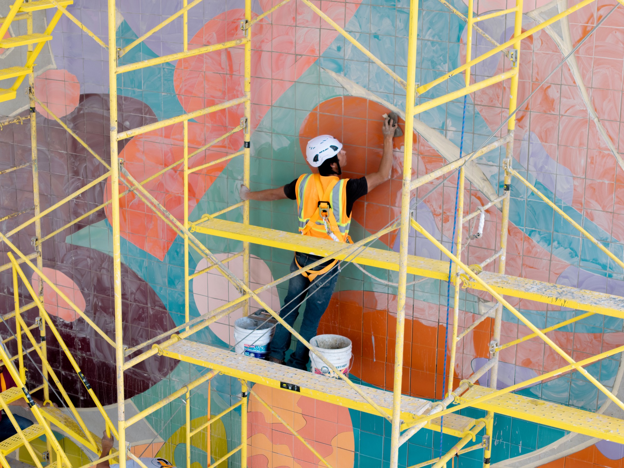 a person works on a mural by Alex Proba of colorful ceramic tiles amid scaffolding
