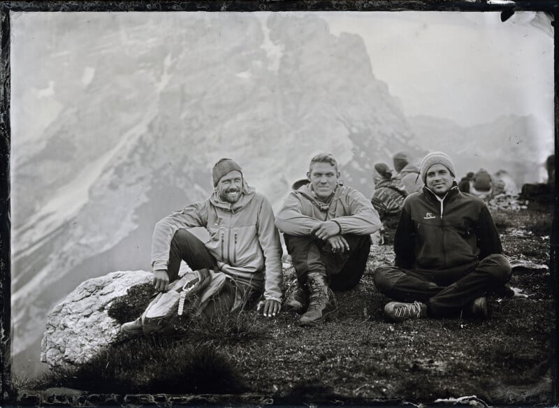 Three people wearing outdoor clothing and beanies sit on a grassy mountain slope with rocky terrain. Snowy, rugged peaks rise in the background. More people are seated in the distance, enjoying the mountainous landscape.