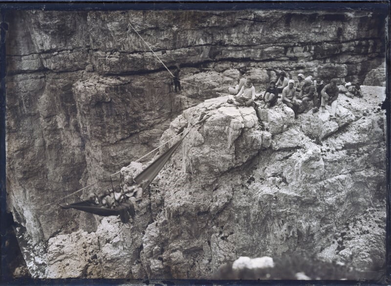 A group of people sits on a rocky cliff while others ride in a suspended hammock-like carrier over a deep canyon, attached to a rope system spanning the gap.