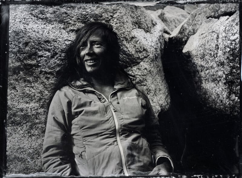 A woman in a Patagonia jacket smiles while sitting against large rocks outdoors. The black-and-white photo has a candid, natural feel.