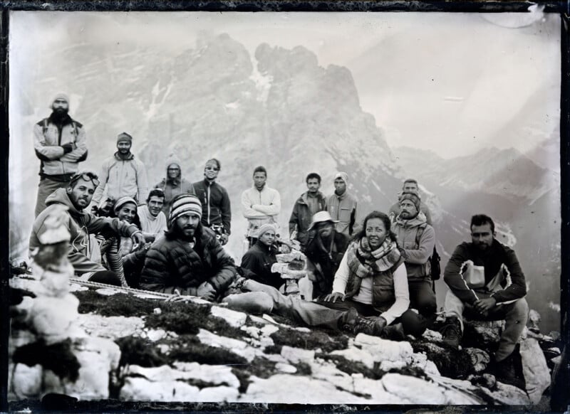 A group of people dressed in outdoor gear sit and stand on rocky ground in a mountainous area, posing for a photo with misty peaks in the background. The image appears to be black and white or sepia-toned.