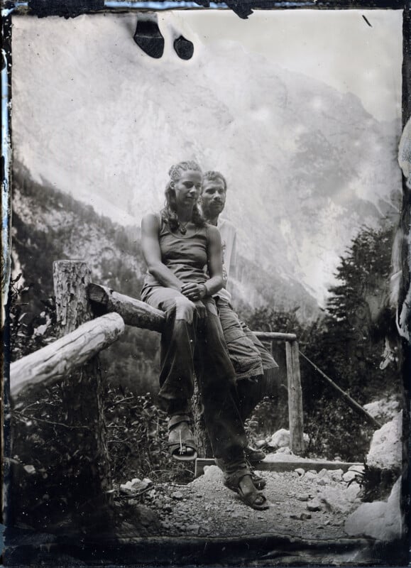 Two people sit closely together on a rustic wooden fence along a mountain trail, surrounded by trees and misty mountains. The photo is in black and white, with a vintage, weathered appearance.