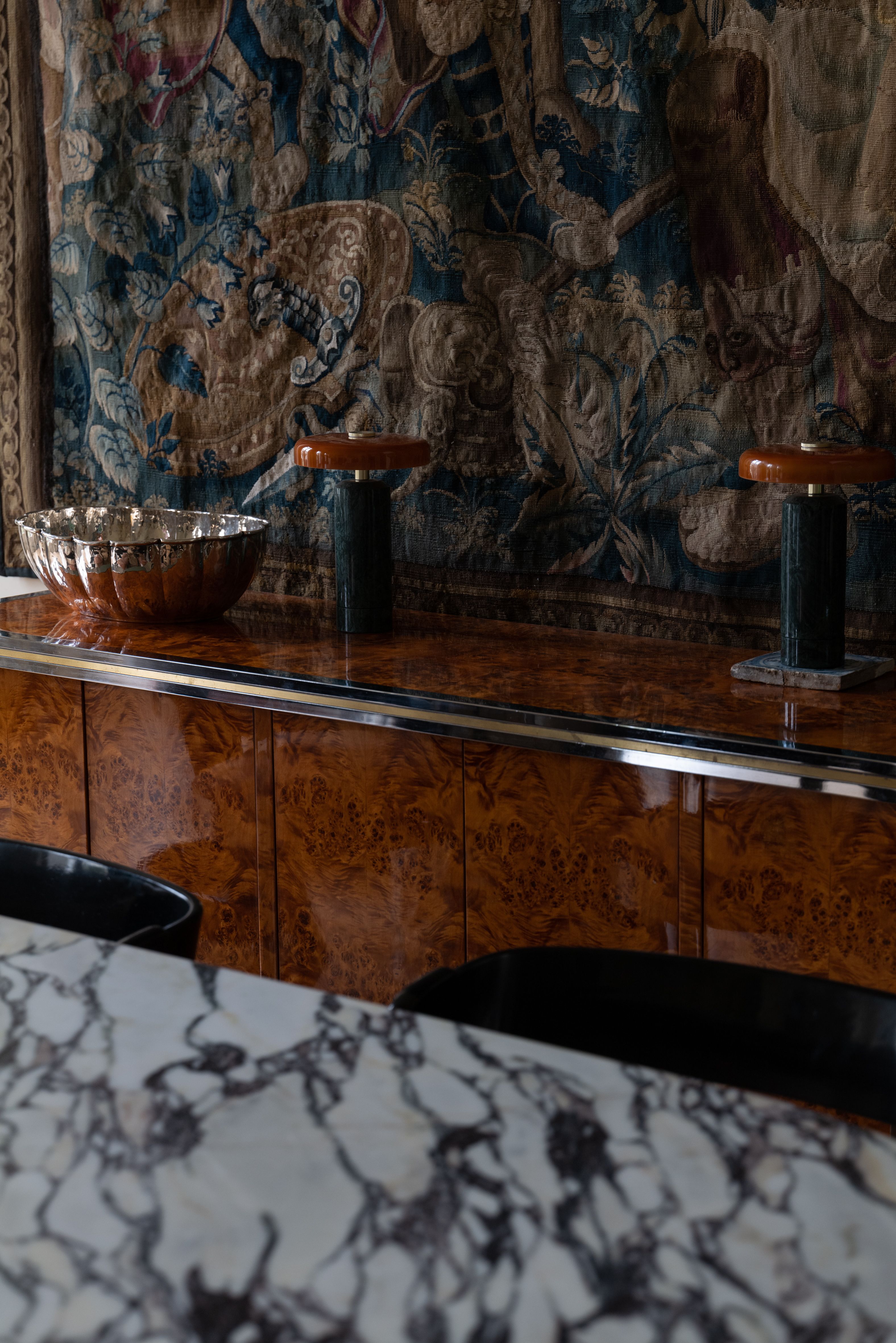 A burl console table below a blue-toned tapestry with two table lamps and a silver bowl on top behind a marble counter