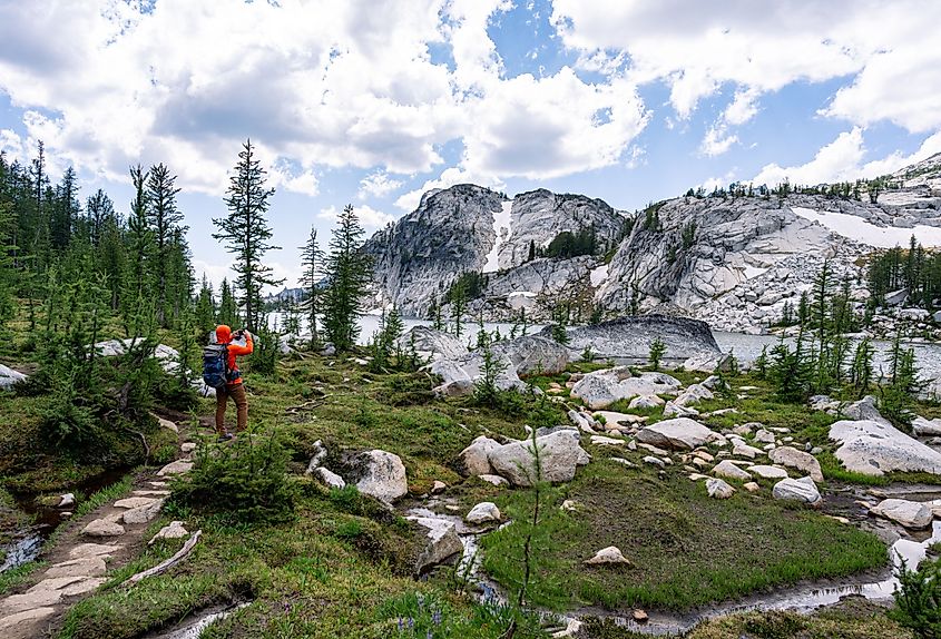 A man in bright orange shirt standing on hiking trail to take picture of beautiful lake and mountain while backpacking in the Enchantments, Leavenworth, Washington