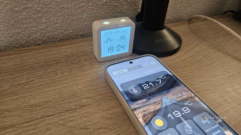 A phone displaying the inside temperature being 21.7 degrees Celsius next to a thermostat showing 25.3 degrees Celsius.