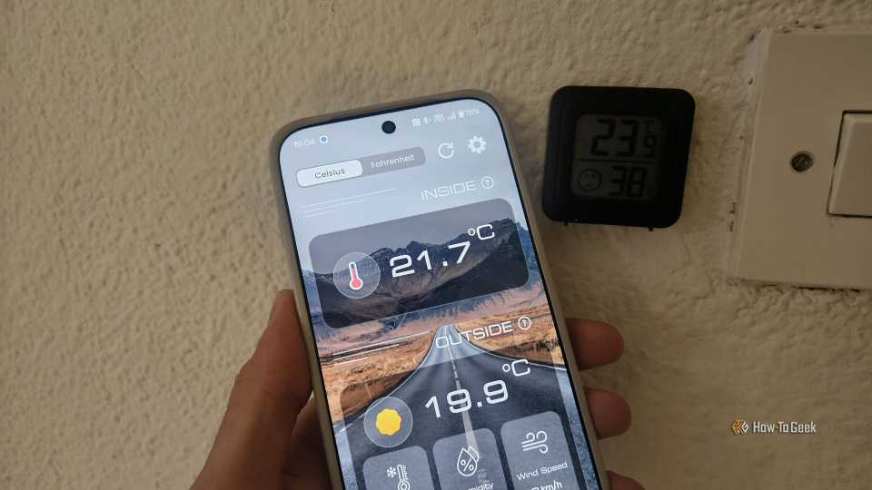 A phone displaying the inside temperature being 21.7 degrees Celsius next to a thermostat showing 23.9 degrees Celsius.