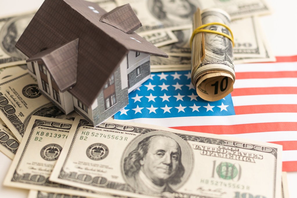 A toy house, rolled-up hundred dollar bills, and scattered hundred dollar bills sit on top of an American flag.
