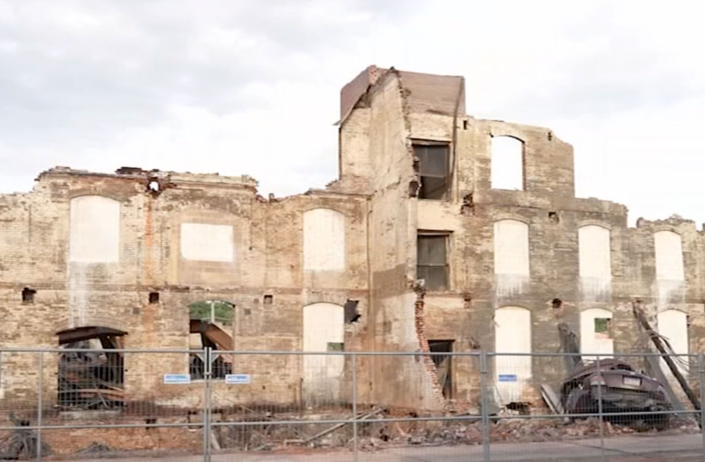 The remains of a severely damaged brick commercial building with a collapsed roof and multiple broken windows, secured by a metal fence.