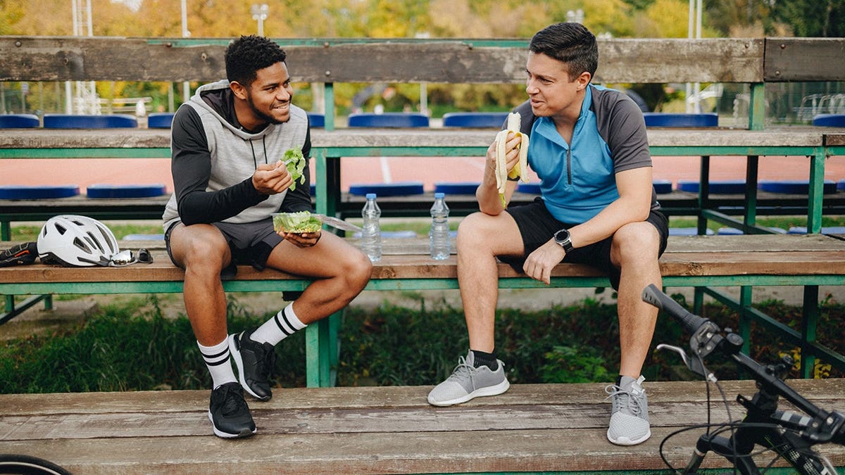 Two young male cyclists sitting on a park bench talking and eating.