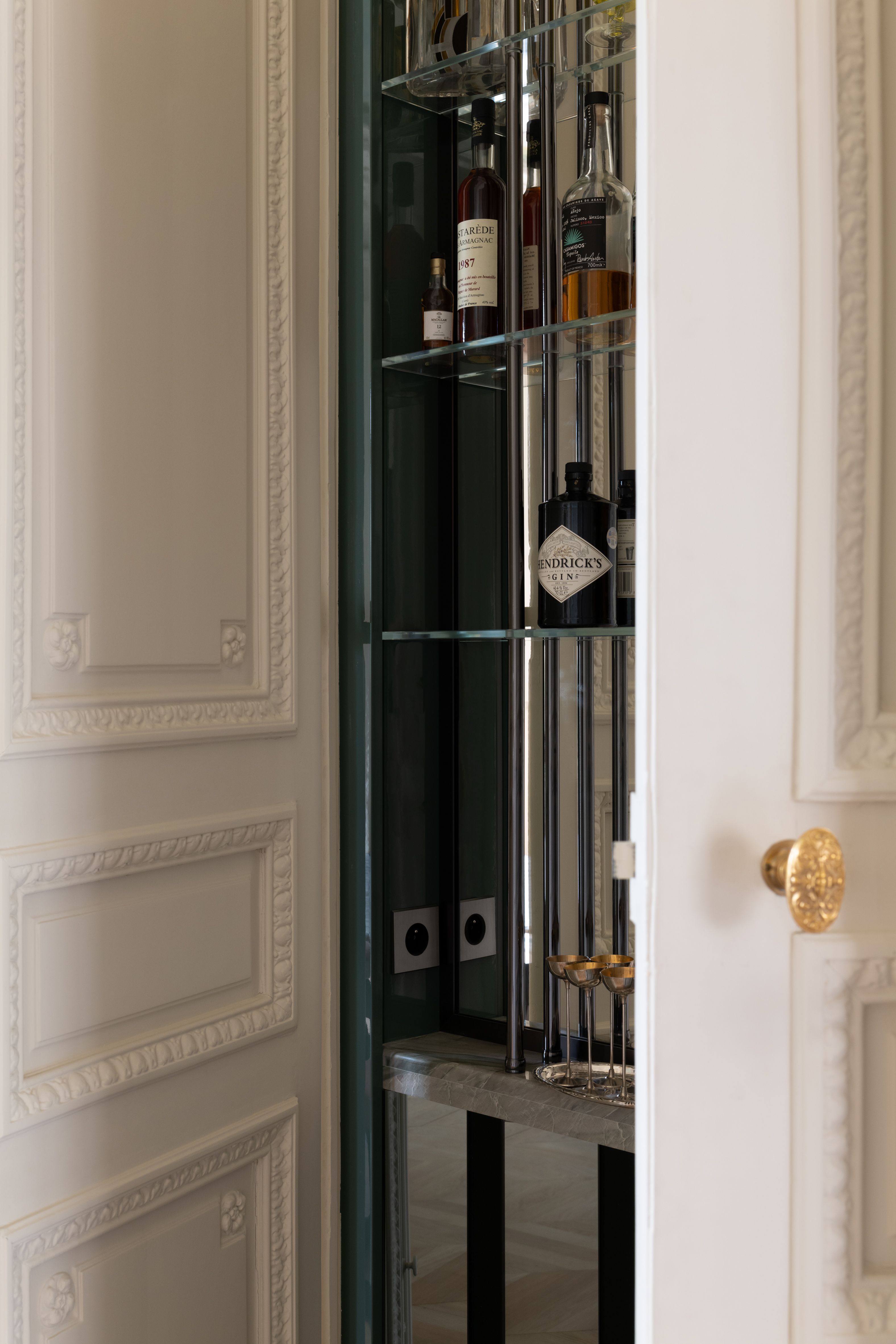 A hidden bar with glass shelving behind an ornate off-white door with gold knobs