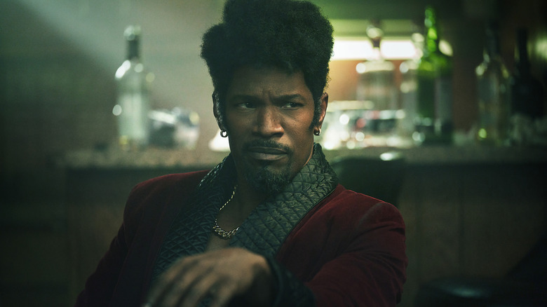 Jamie Foxx as Slick Charles sitting at a table with a drink in They Cloned Tyrone