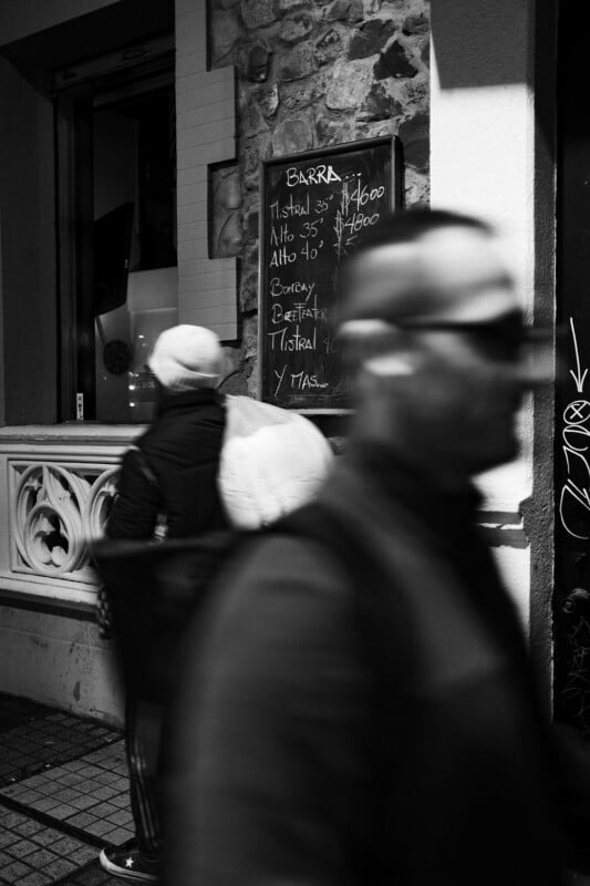 Black and white photo of two people walking past a stone building with a chalkboard menu listing drinks in Spanish. One person is blurred in the foreground; the other wears a hat and backpack. Urban street scene at night.