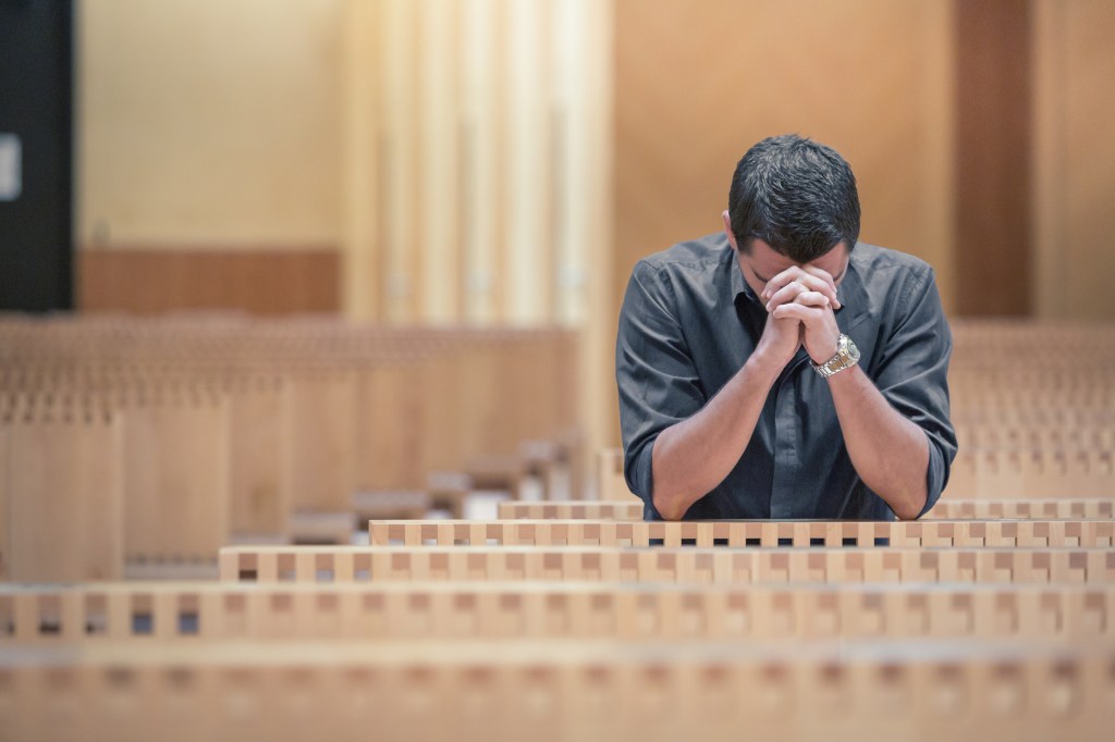 Young man with a beard, wearing a blue shirt, praying in a modern church with his hands clasped together.