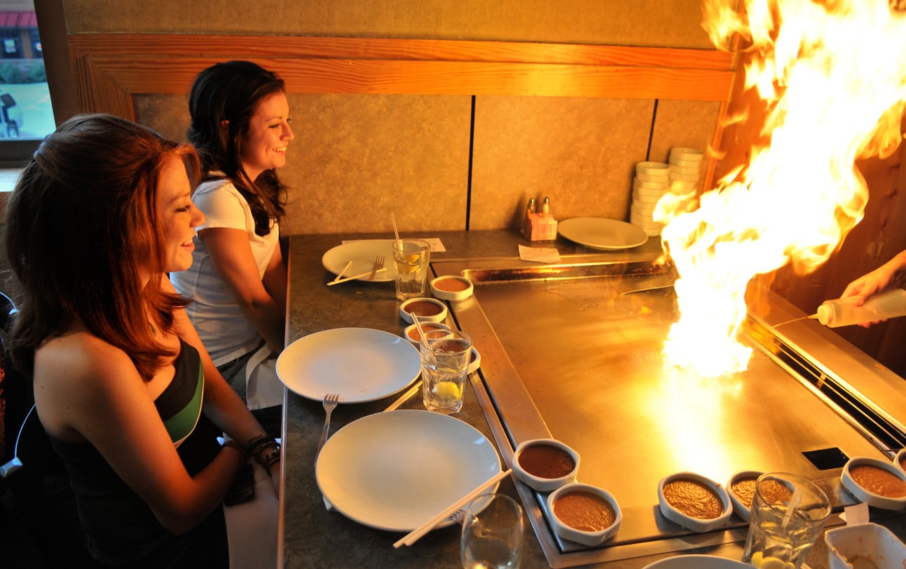 Two women sit at a hibachi grill table, smiling as a chef creates a large flame on the cooking surface—a signature experience at one of the city's popular tourist attractions. Plates, drinks, and dipping sauces are arranged in front of them.