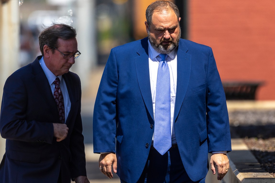 Two balding men wearing suits walk side by side outdoors. The man on the right wears a blue suit, a blue tie and rings on both ring fingers.