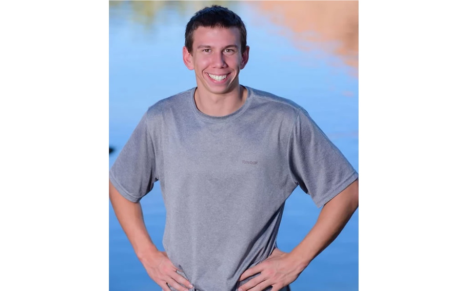 A young man in a gray T-shirt smiles for a portrait outdoors. 