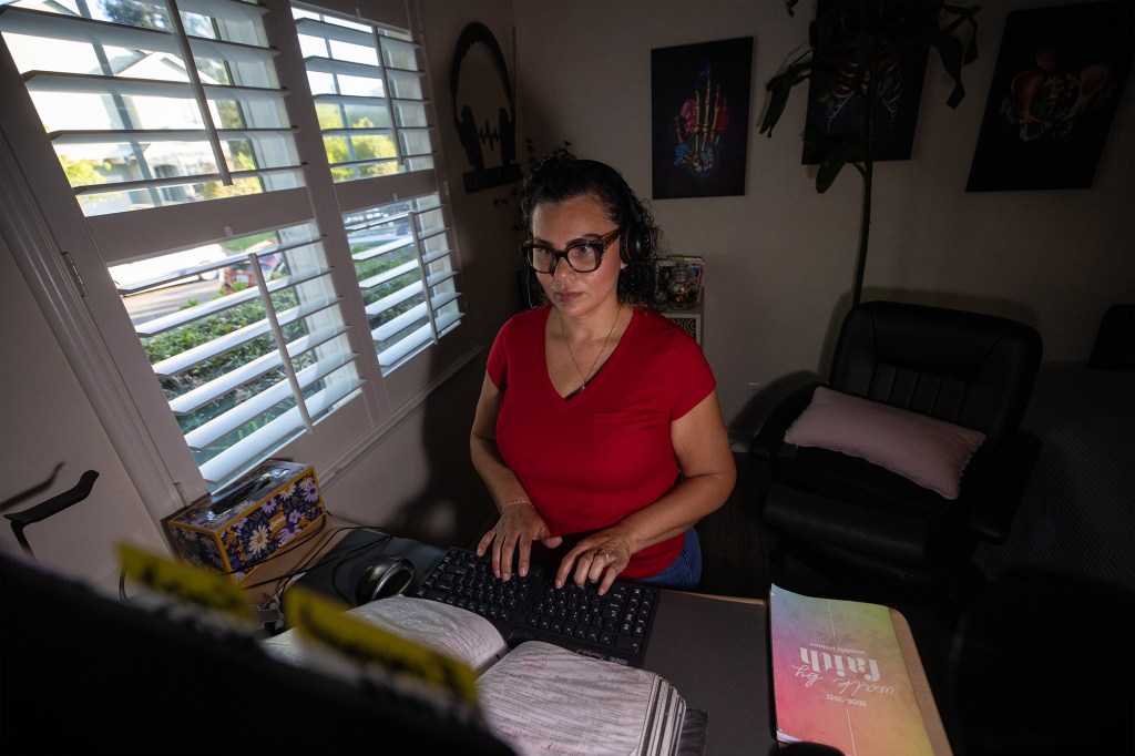 A person wearing a red shirt and a headset sits in front of a desk and types into a keyboard, in a dimly lit room with a window overlooking a residential street.