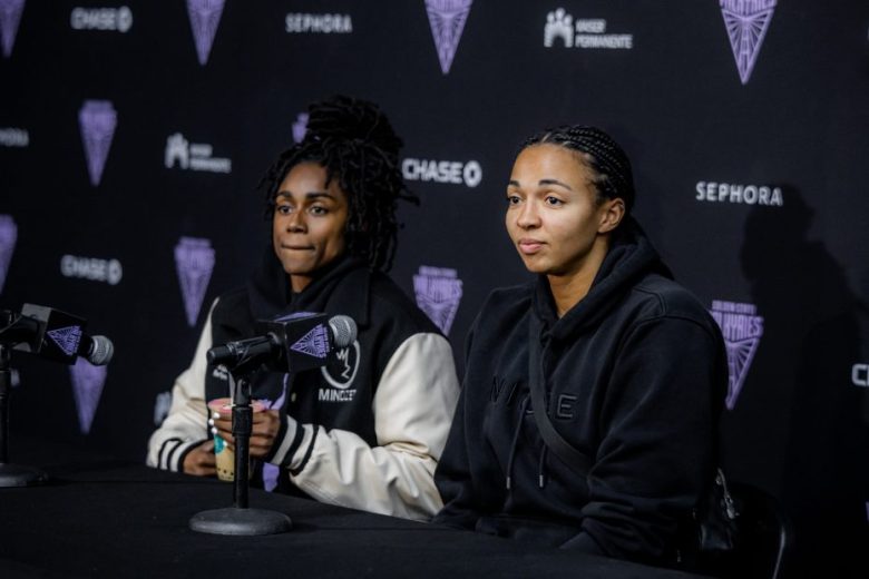 Two women sit at a press conference table with microphones in front of a backdrop featuring various sponsor logos.