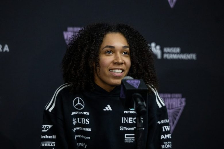 A person with curly hair speaks at a press conference, wearing a black sweatshirt with multiple sponsor logos against a branded backdrop.