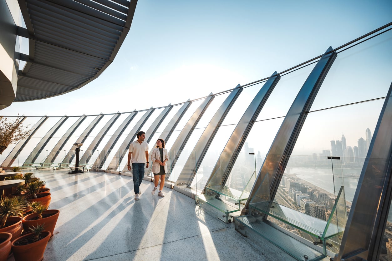 A couple walks along a modern glass observation deck overlooking a city skyline on a sunny day, with potted plants lining the walkway—one of the city's most popular tourist attractions.