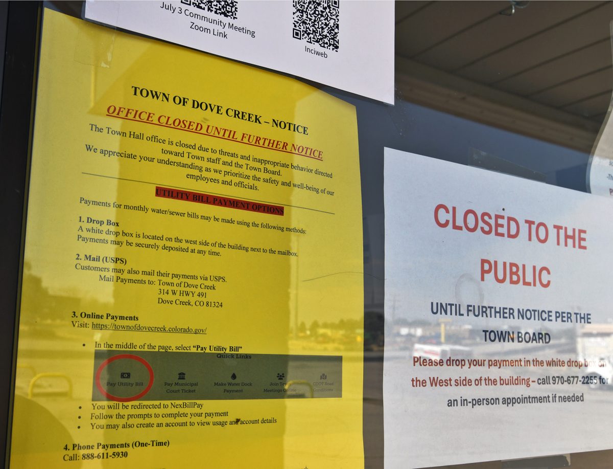 Signs on the door of Dove Creek's town hal warn visitors that the office is closed until further notice. The yellow signs explain how people can pay their utility bills by dropping check in a box, mailing, or paying online. The white sign to the left tells people they can pay in person if they make an appointment. And the white sign above the yellow sign has QR codes directing visitors to information about a town meeting on the Ferris fire and to a website with fire information