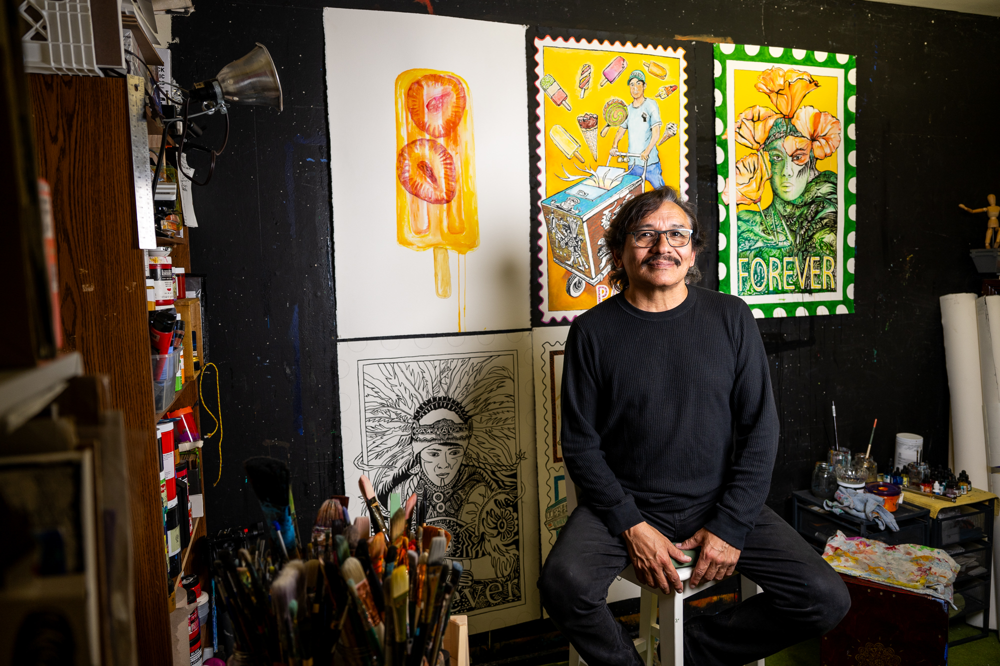 man in glasses on stool in painting studio with artworks behind him