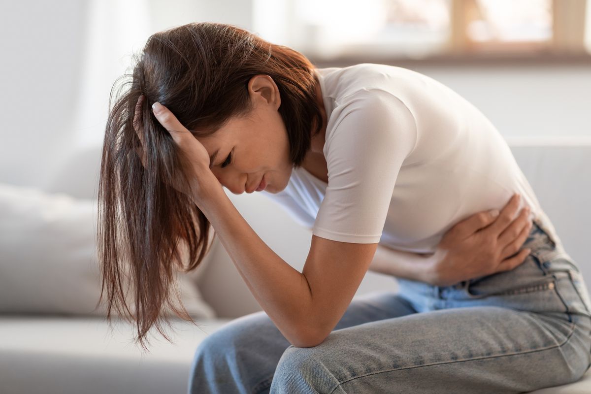 Young lady touching her stomach and head in distress, suffering from headache and stomachache, having symptoms of PMS and gastrointestinal discomfort, sitting in living room at home