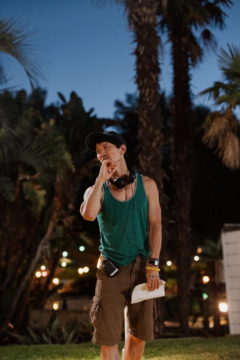 A person wearing headphones and a tank top stands outdoors, holding a sheet of paper and thinking.