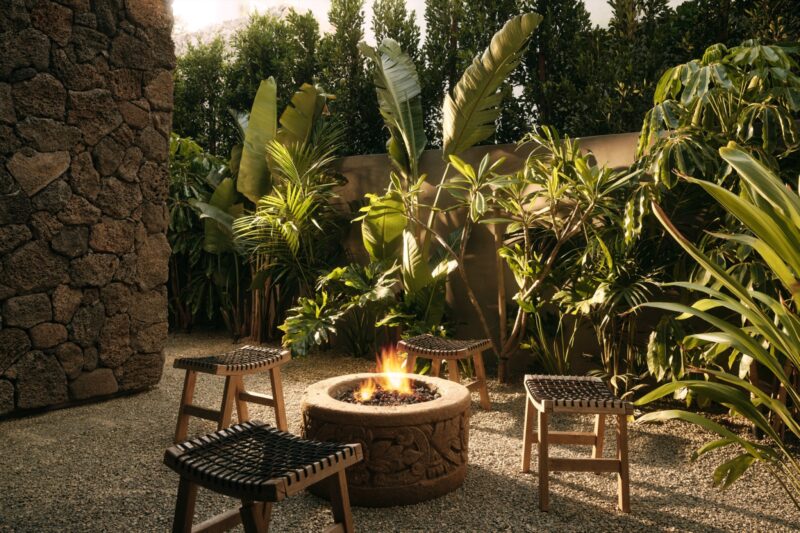 Four woven stools surround a stone fire pit with a small fire, set in the gravel courtyard of Obsidian House, framed by dense tropical plants and a striking stone wall.