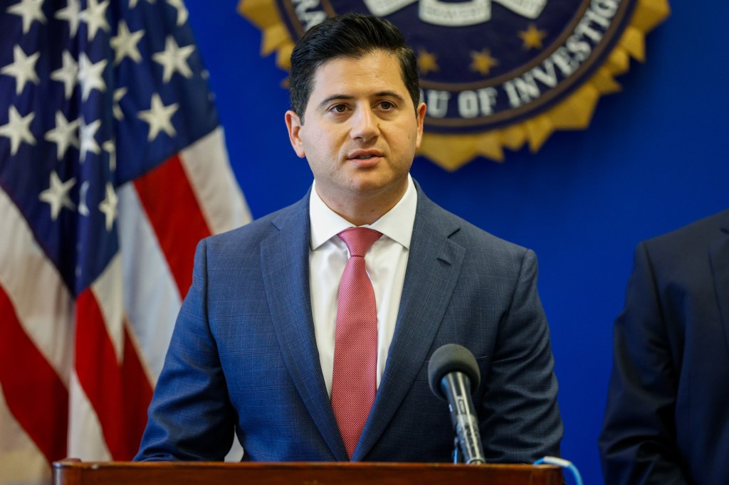 A man in a suit speaks at a podium with an American flag and a seal in the background.