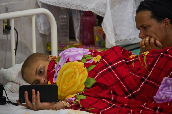 Salet Fernandez, a 17-year-old oncology patient, looks at her phone as her mother Yarima Mesa sits by her bed at the National Institute of Oncology and Radiobiology in Havana, Cuba, Wednesday, June 24, 2026. (AP Photo/Ramon Espinosa)