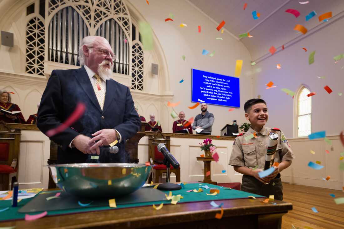 The Rev. John Jackman, a man with a white beard and hair, and a boy of about 9 in a Scout uniform are shown on the altar of a church, with colorful confetti floating around them. 