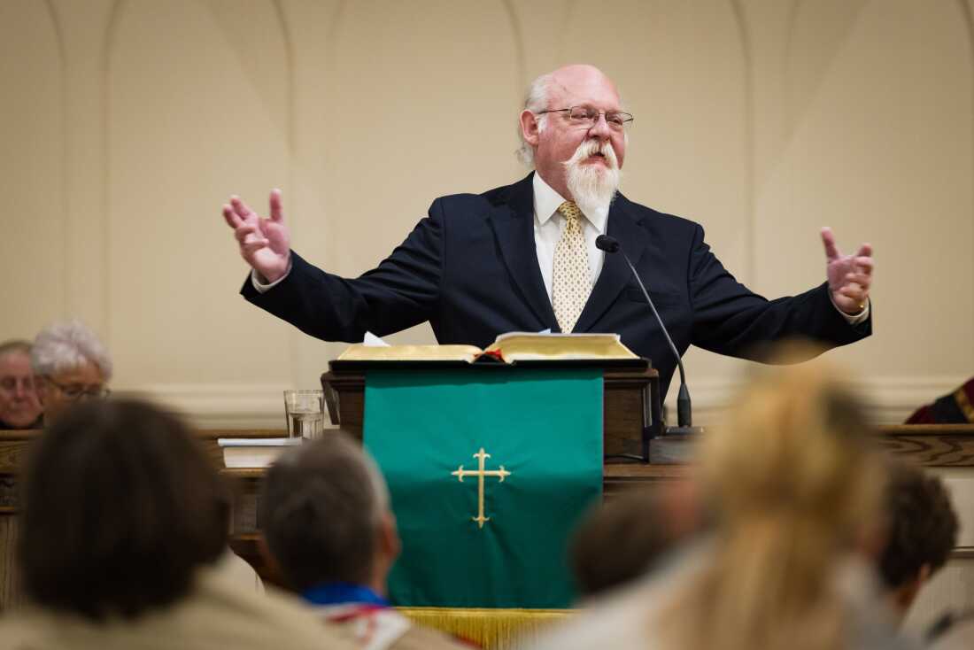 The Rev. John Jackman is standing at a pulpit, speaking. His arms are open wide.