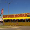 A bright yellow fireworks stand is under a clear blue sky with a large sign at the top of the stand the that reads "Buy one, get one free!" in large bold, capitol red letters. The word 'Fireworks,' is to the right of it in larger bold, capitol red letters. Red, white and blue fringe hangs from the awning. Red flags with white stars are attached on either side of the stand.