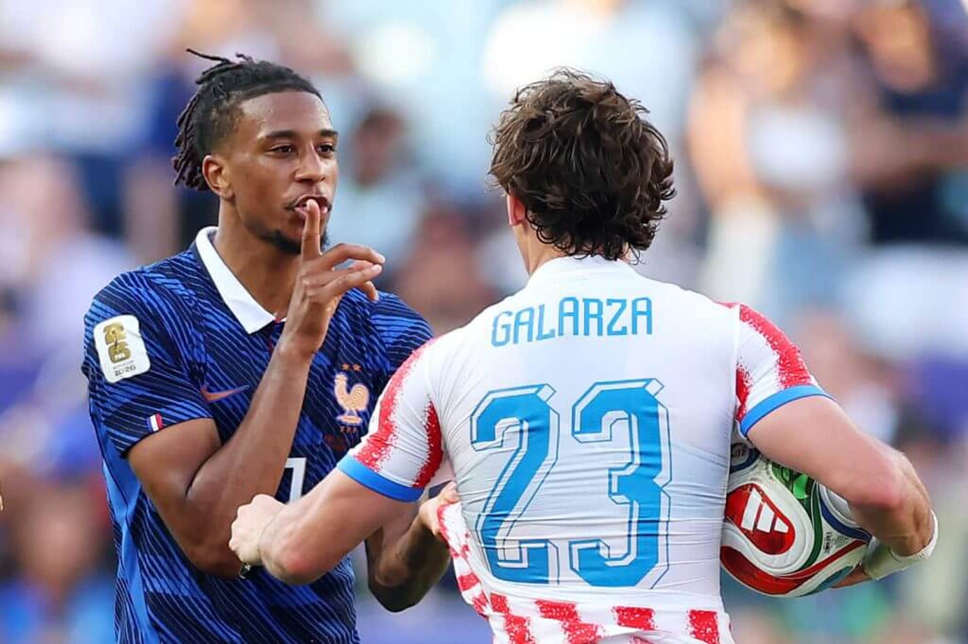 Michael Olise holds his finger over his mouth in a 'shushing' gesture during a confrontation with Paraguay's Matias Galarza, whose back is to the camera.