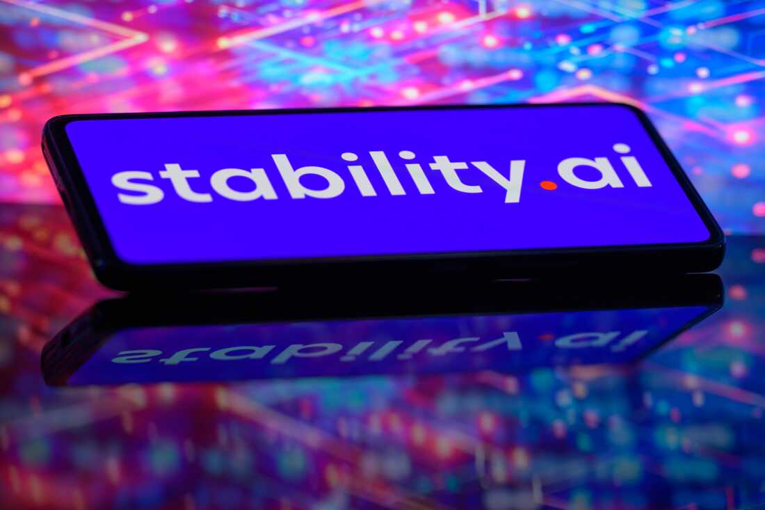 The Stability AI logo is displayed on a smartphone screen. The screen, which is tilted backwards at an angle, is blue and the company logo is white and red. The smart phone is shown against a blue and red abstract background.