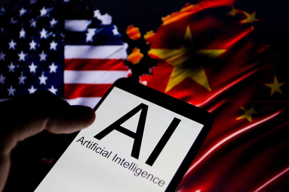 This photo is a close-up view of a smartphone whose screen is displaying "AI" in large letters and the words "Artificial Intelligence" underneath. In the background are the blurred flags of the United States (on the left) and China (on the right).