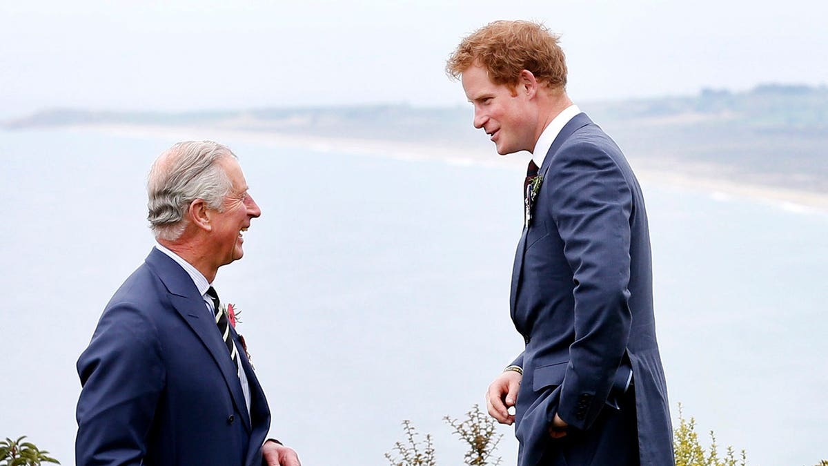 King Charles and a younger Prince Harry wearing matching dark blue suits in conversation and smiling.