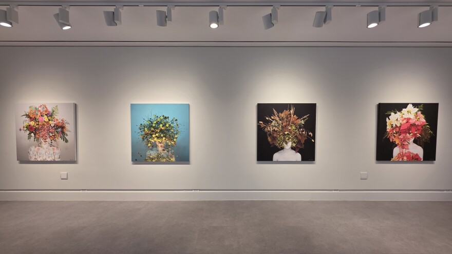 Four paintings hang on a museum wall depicting human figures with giant flower headdresses on.