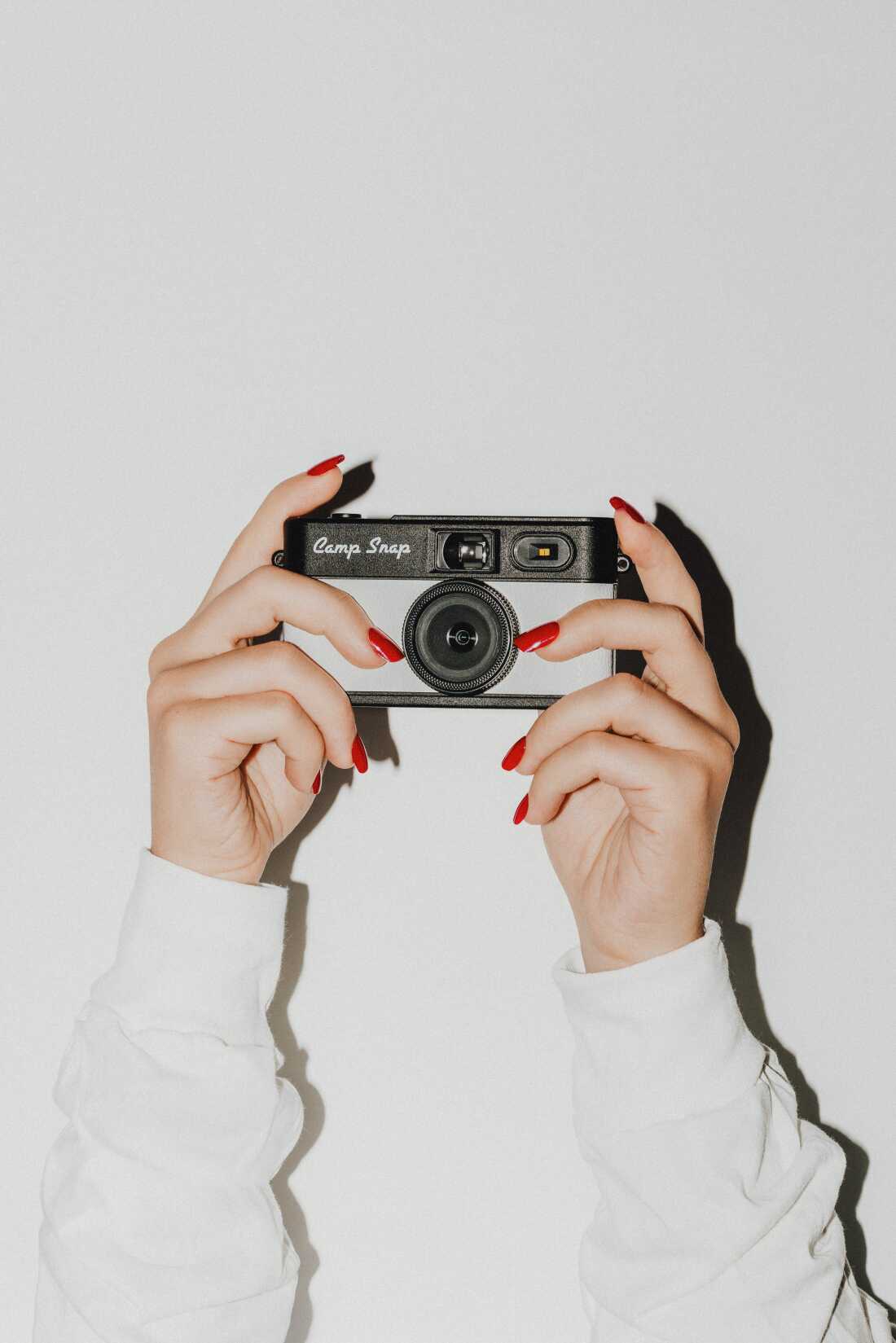This photo shows a beige and black Camp Snap camera that is being held by a woman's hands, which have long red-pained fingernails.