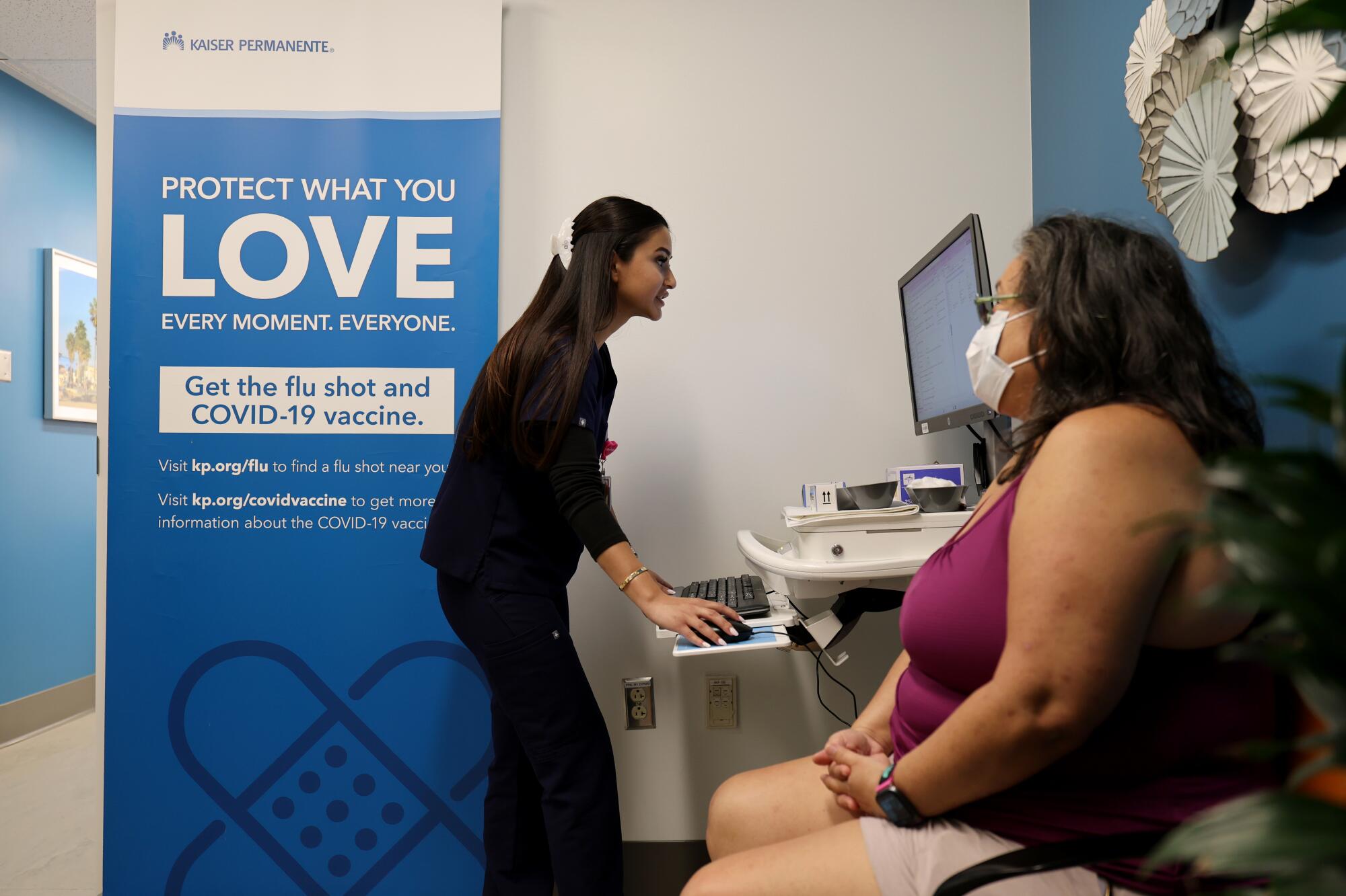 A nurse enters data into a computer at a standing desk while a patient with a mask sits nearby