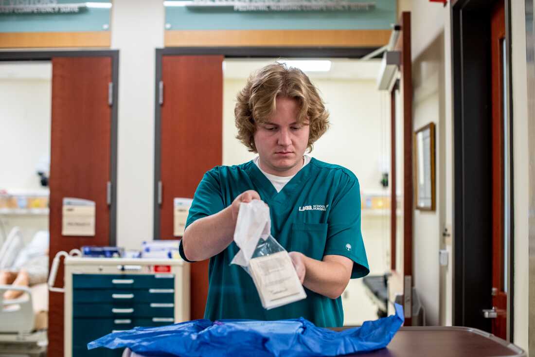 Nursing student Ethan Stinson prepares personal protective equipment during a class exercise. He has blonde curly hair and is wearing teal scrubs.