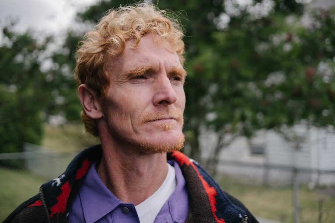 Cody Coughenour stands for a head-and-shoulders portrait outside his home. He has curly red hair and a determined expression on his face.